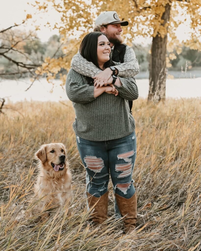 A young couple poses in the trees by the river with their Golden Retriever.